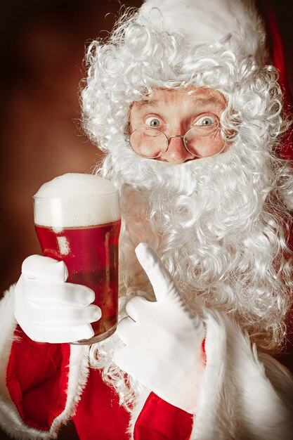 Porträt des Mannes im Weihnachtsmannkostüm mit einem luxuriösen weißen Bart, der Weihnachtsmannmütze und einem roten Kostüm auf Rot mit Bier
