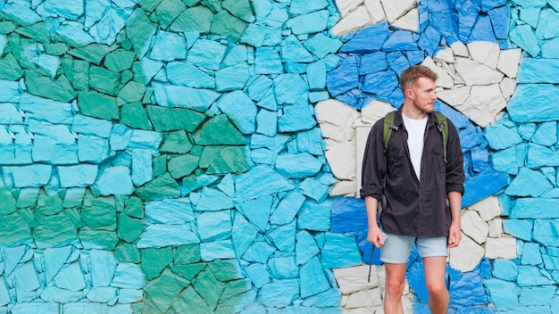 Kostenloses Foto porträt des mannes mit dem rucksack, der gegen gemalte steinwand steht