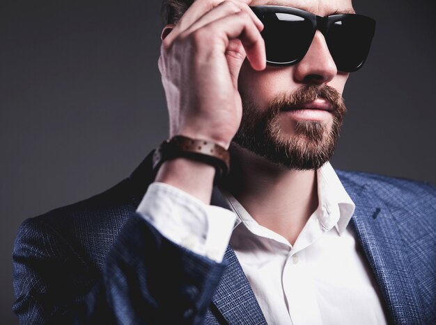 Porträt des modischen Hipster-Geschäftsmannmodells der schönen Mode, gekleidet im eleganten blauen Anzug, der auf Grau aufwirft