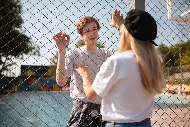 Porträt des nachdenklichen Jungen, der auf dem Basketballplatz steht und hübsches Mädchen mit blonden Haaren durch Maschendrahtzaun verträumt betrachtet Junges schönes Paar, das steht und einander anschaut