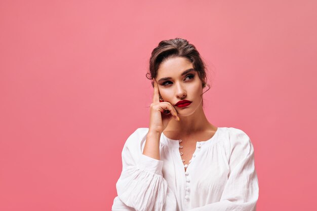 Porträt des nachdenklichen Mädchens mit den roten Lippen auf rosa Hintergrund. Ernsthafte junge Frau in der weißen stilvollen Bluse, die auf Kamera aufwirft.