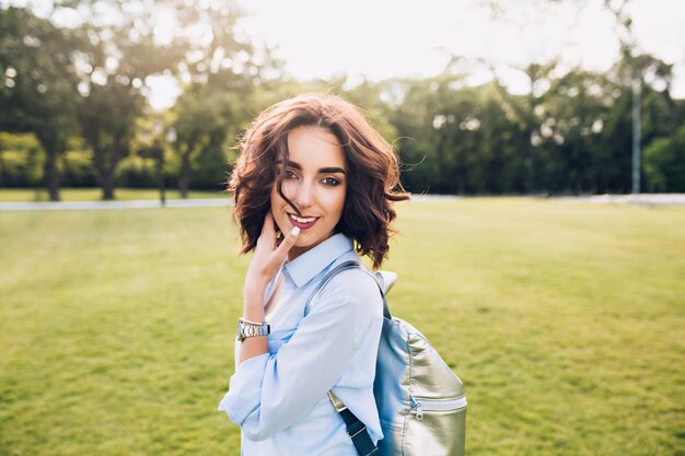 Porträt des netten Brunettemädchens mit dem kurzen Haar, das auf Sonnenuntergang im Park geht. Sie trägt blaues Hemd und Tasche. Sie lächelt in die Kamera.