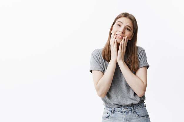 Porträt des niedlichen kaukasischen Studentenmädchens mit dunklem Haar und braunen Augen im grauen T-Shirt und in den Jeans, die sanft lächeln, mit weichem und entspanntem Blick, Gesicht mit den Fingern berührend