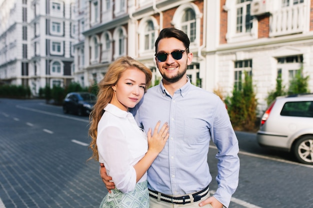 Porträt des niedlichen Paares, das um britisches Viertel geht. Hübscher Kerl in der Sonnenbrille, die blondes Mädchen mit langen Haaren umarmt und lächelt, sie verliert ernsthaft.