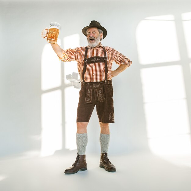 Porträt des Oktoberfest-älteren Mannes im Hut, der die traditionelle bayerische Kleidung trägt. Männlicher Schuss in voller Länge im Studio auf weißem Hintergrund. Die Feier, Feiertage, Festivalkonzept. Bier trinken.