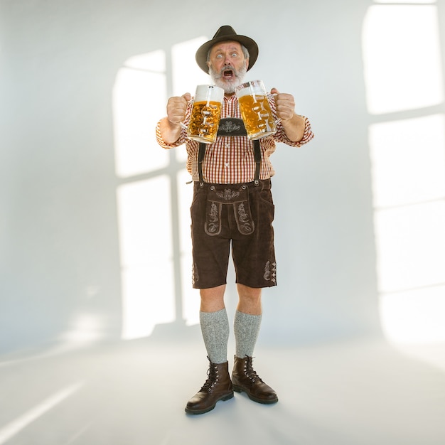 Kostenloses Foto porträt des oktoberfest-älteren mannes im hut, der die traditionelle bayerische kleidung trägt. männlicher schuss in voller länge im studio auf weißem hintergrund. die feier, feiertage, festivalkonzept. bier trinken.