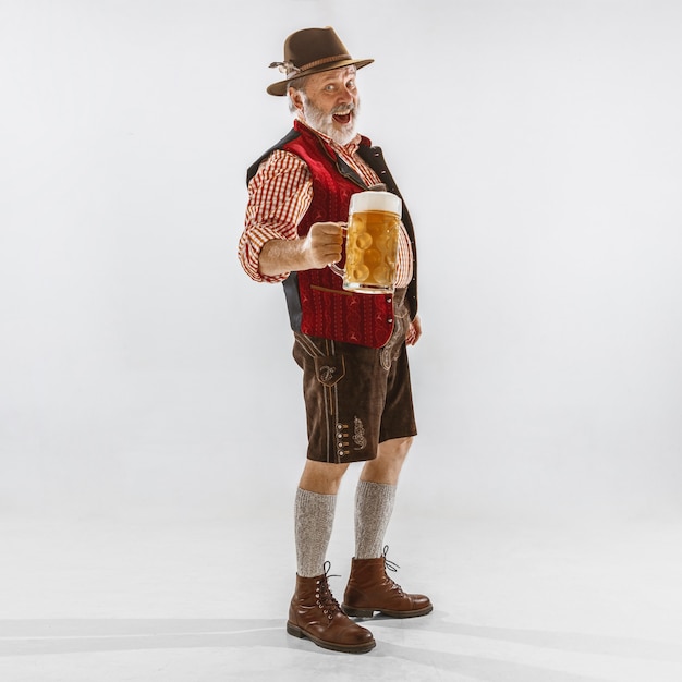 Porträt des Oktoberfest-älteren Mannes im Hut, der die traditionelle bayerische Kleidung trägt. Männlicher Schuss in voller Länge im Studio auf weißem Hintergrund. Die Feier, Feiertage, Festivalkonzept. Bier trinken.
