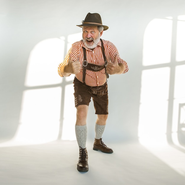 Porträt des Oktoberfest-älteren Mannes im Hut, der die traditionelle bayerische Kleidung trägt. Männlicher Schuss in voller Länge im Studio auf weißem Hintergrund. Die Feier, Feiertage, Festivalkonzept. Geste nett.