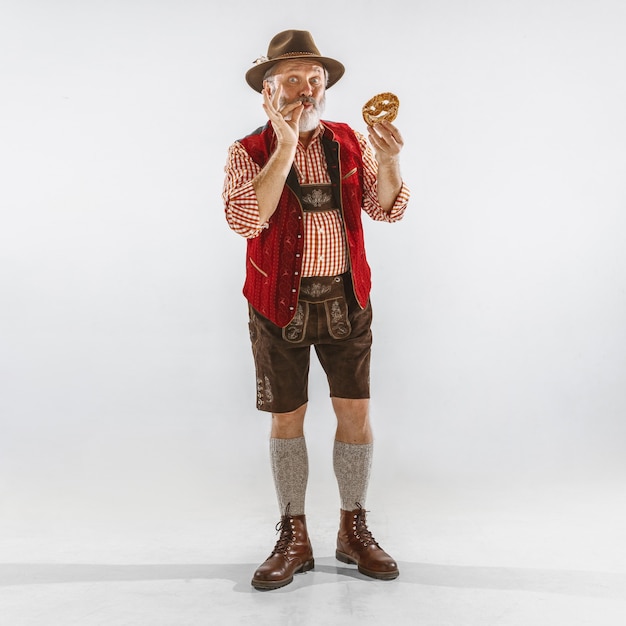Porträt des Oktoberfest-älteren Mannes im Hut, der die traditionelle bayerische Kleidung trägt. Männlicher Schuss in voller Länge im Studio auf weißem Hintergrund. Die Feier, Feiertage, Festivalkonzept. Puff essen.