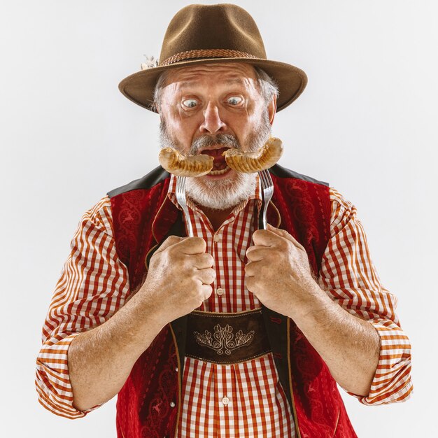Porträt des Oktoberfest-älteren Mannes im Hut, der die traditionelle bayerische Kleidung trägt. Männlicher Schuss in voller Länge im Studio auf weißem Hintergrund. Die Feier, Feiertage, Festivalkonzept. Würstchen essen.