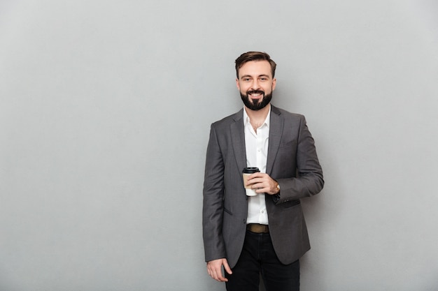 Porträt des positiven Mannes im weißen Hemd, das auf Kamera mit breitem Lächeln aufwirft und Mitnehmerkaffee über Grau hält