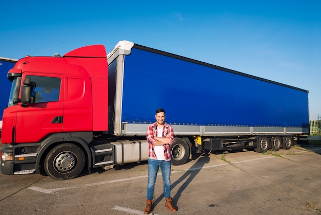 Porträt des professionellen amerikanischen LKW-Fahrers in der Freizeitkleidung und in den Stiefeln, die vor LKW-Fahrzeug mit langem Anhänger stehen