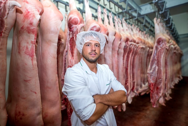 Porträt des professionellen Metzgers im Fabrikkühlhaus, das Arme hält, gekreuzt mit Schweineschlachtkörper im Hintergrund