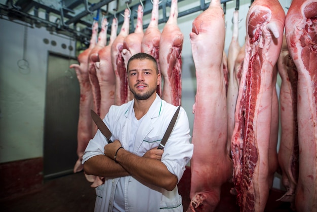 Porträt des professionellen Metzgers im Fabrikkühlraum, der scharfe Messer mit Schweineschlachtkörper dahinter hält