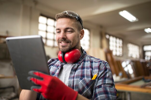 Porträt des professionellen Zimmermanns mittleren Alters mit Gehörschutz unter Verwendung der Tablette in der Tischlerwerkstatt