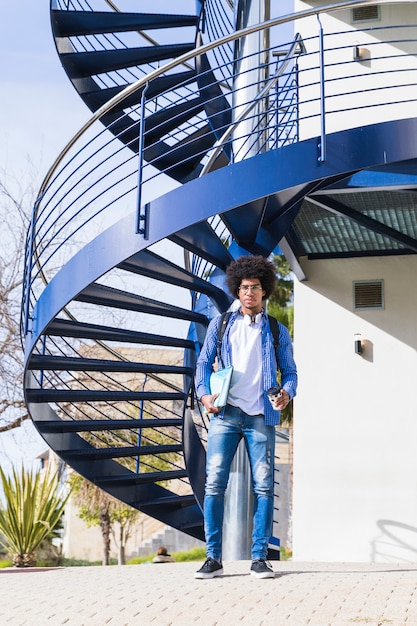 Porträt des reizend hübschen männlichen Studenten der Universität, der vor blauem Wendeltreppe steht