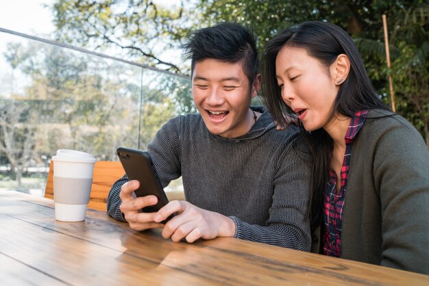 Porträt des reizenden asiatischen Paares, das das Handy beim Sitzen und Verweilen im Café betrachtet.