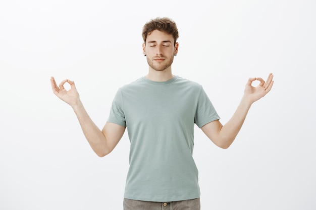 Porträt des ruhigen gutaussehenden kaukasischen Mannes im T-Shirt, der lächelt und sich entspannt fühlt, mit gespreizten Händen in Zen-Geste und geschlossenen Augen stehend