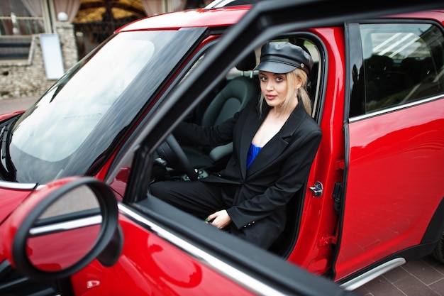 Porträt des schönen blonden sexy Modefrauenmodells in der Kappe und ganz in Schwarz mit hellem Make-up sitzen und fahren rotes Stadtauto