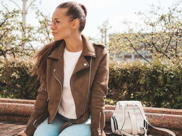 Porträt des schönen Brunettemodells kleidete in der Sommerhippie-Jacke und in der Jeanskleidung an Modisches Mädchen, das auf der Bank in der Straße sitzt