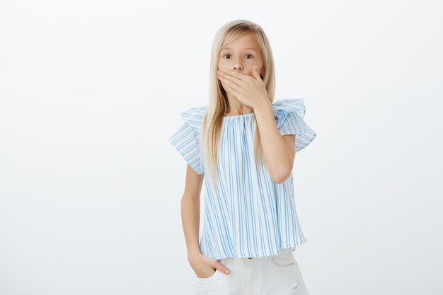 Porträt des schönen geschockten Mädchens mit blondem Haar in der blauen Bluse, keuchend, den Mund bedeckend, um nicht vor Angst zu schreien, mit gruseliger Spinne erstaunt und erschrocken, gegen graue Wand stehend