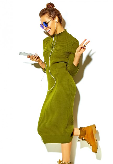 Porträt des schönen glücklichen niedlichen lächelnden brünetten Frauenmädchens in der lässigen grünen Hipster-Sommerkleidung lokalisiert auf Weiß in der Sonnenbrille, die Musik im Smartphone mit Kopfhörern hört