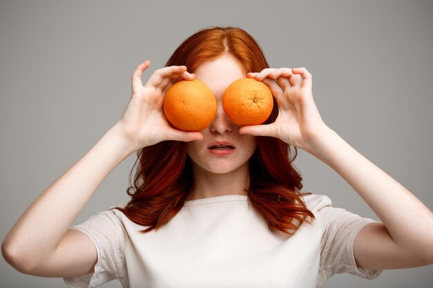 Porträt des schönen Ingwermädchens, das Orangen über grauem Hintergrund hält