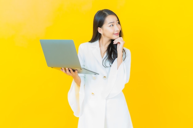 Porträt des schönen jungen asiatischen Frauenlächelns mit Computerlaptop auf gelber Wand