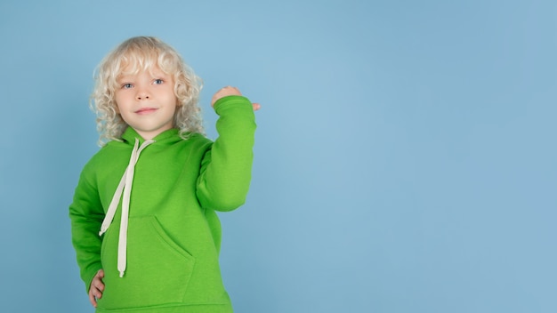 Porträt des schönen kleinen kaukasischen Jungen lokalisiert auf blauem Studiohintergrund. Blondes lockiges männliches Model. Konzept des Gesichtsausdrucks, der menschlichen Emotionen, der Kindheit, der Anzeige, des Verkaufs.