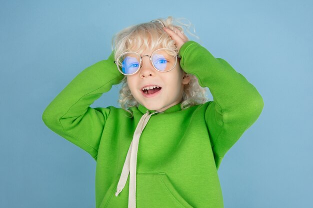 Porträt des schönen kleinen kaukasischen Jungen lokalisiert auf blauem Studiohintergrund. Blondes lockiges männliches Model. Konzept des Gesichtsausdrucks, der menschlichen Emotionen, der Kindheit, der Anzeige, des Verkaufs.