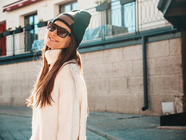 Porträt des schönen lächelnden Baumusters. Frau gekleidet in warmen Hipster-weißen Pullover und Mütze. Trendiges Mädchen posiert auf der Straße
