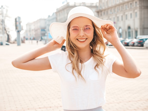 Porträt des schönen lächelnden blonden Modells kleidete in der Sommerhippie-Kleidung an. Modisches Mädchen, das in der Straße in der runden Sonnenbrille aufwirft. Lustige und positive Frau, die Spaß im Hut hat