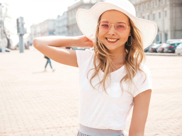 Porträt des schönen lächelnden blonden Modells kleidete in der Sommerhippie-Kleidung an. Modisches Mädchen, das in der Straße in der runden Sonnenbrille und im Hut aufwirft. Lustige und positive Frau, die Spaß hat