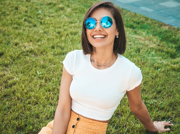 Porträt des schönen lächelnden brünetten Modells gekleidet in Sommerhipperkleidung. Trendy Mädchen, das auf dem Gras im Park sitzt. Lustige und positive Frau, die Spaß hat