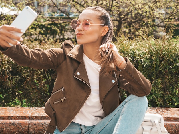 Porträt des schönen lächelnden Brunettemädchens in der Sommerhippie-Jacke und -jeans modellieren das Nehmen von selfie auf Smartphone