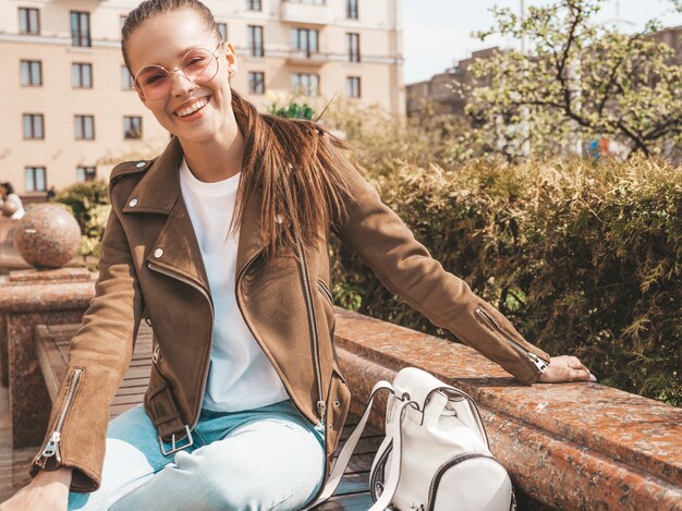 Porträt des schönen lächelnden Brunettemodells kleidete in der Sommerhippie-Jacke und in der Jeanskleidung an Modisches Mädchen, das auf der Bank in der Straße sitzt