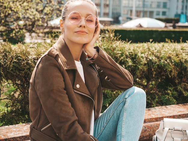 Porträt des schönen lächelnden Brunettemodells kleidete in der Sommerhippie-Jacke und in der Jeanskleidung an Modisches Mädchen, das auf der Bank in der Straße sitzt