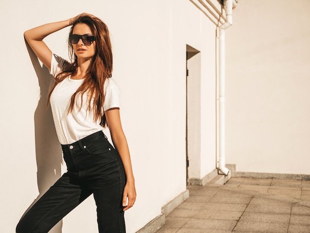 Porträt des schönen lächelnden Modells in der Sonnenbrille. Frau gekleidet im Sommer Hipster weißes T-Shirt und Jeans.Posing in der Nähe der Wand auf der Straße