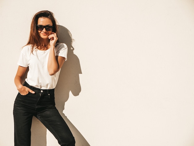 Porträt des schönen lächelnden Modells in der Sonnenbrille. Frau gekleidet im Sommer Hipster weißes T-Shirt und Jeans.Posing in der Nähe der Wand auf der Straße