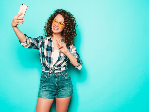 Porträt des schönen lächelnden Modells mit Afro-Lockenfrisur gekleidet in Sommer-Hipster-Kleidung. Sexy sorgloses Mädchen, das im Studio auf grauem Hintergrund aufwirft. Trendige lustige Frau macht Selfie-Foto