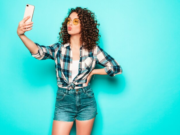Porträt des schönen lächelnden Modells mit Afro-Lockenfrisur gekleidet in Sommer-Hipster-Kleidung. Sexy sorgloses Mädchen, das im Studio auf grauem Hintergrund aufwirft. Trendige lustige Frau macht Selfie-Foto