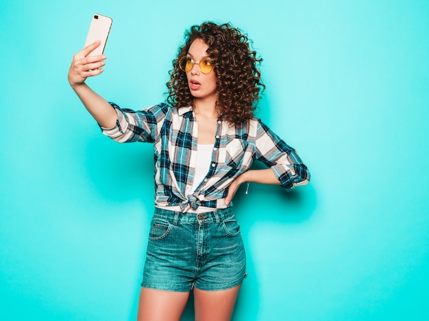 Porträt des schönen lächelnden Modells mit Afro-Lockenfrisur gekleidet in Sommer-Hipster-Kleidung. Sexy sorgloses Mädchen, das im Studio auf grauem Hintergrund aufwirft. Trendige lustige Frau macht Selfie-Foto