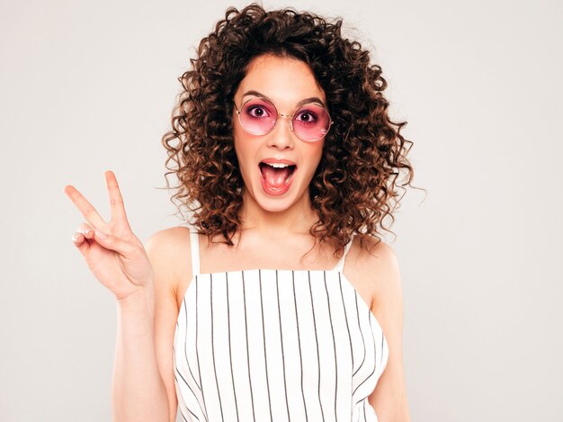 Porträt des schönen lächelnden Modells mit Afro-Lockenfrisur gekleidet in Sommer-Hipster-Kleidung. Trendige lustige und positive Frau zeigt Friedenszeichen