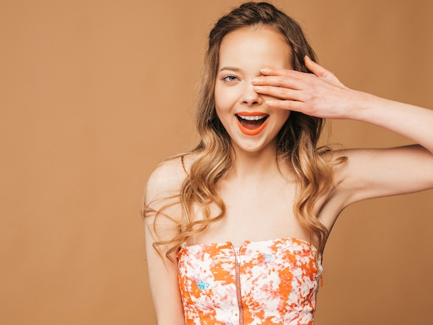 Porträt des schönen lächelnden netten Modells mit den rosa Lippen. Mädchen im bunten Sommerkleid. Model posiert. Bedeckte ihr Auge mit der Hand