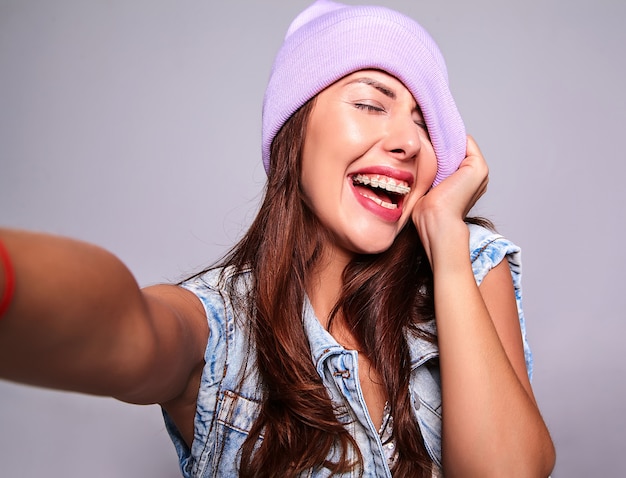 Porträt des schönen lächelnden niedlichen brünetten Frauenmodells in der lässigen Sommerjeanskleidung ohne Make-up in der lila Mütze, die Selfie-Foto am Telefon lokalisiert auf grau macht