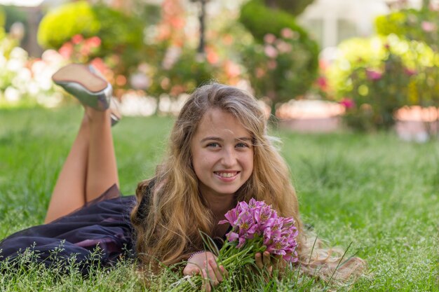 Porträt des schönen Mädchens außerhalb liegend, lächelnd, während Blumen im schwarzen T-Shirt während des Tages halten.