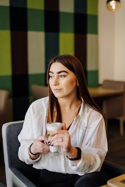 Porträt des schönen Mädchens, das heißen Tee oder Kaffee in einem Café mit ihrem Handy trinkt.