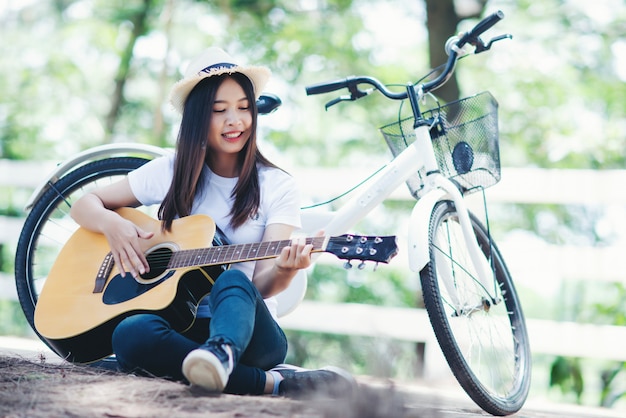 Porträt des schönen Mädchens die Gitarre mit Fahrrad an der Natur spielend