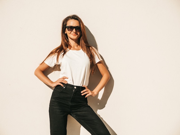 Porträt des schönen Modells in der Sonnenbrille. Frau gekleidet im Sommer Hipster weißes T-Shirt und Jeans. Trendiges Mädchen posiert in der Nähe der Wand auf der Straße