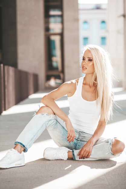 Porträt des schönen netten blonden Mädchens im weißen T-Shirt und in den Jeans, die draußen aufwerfen. Nettes Mädchen, das auf Asphalt auf der Straße sitzt